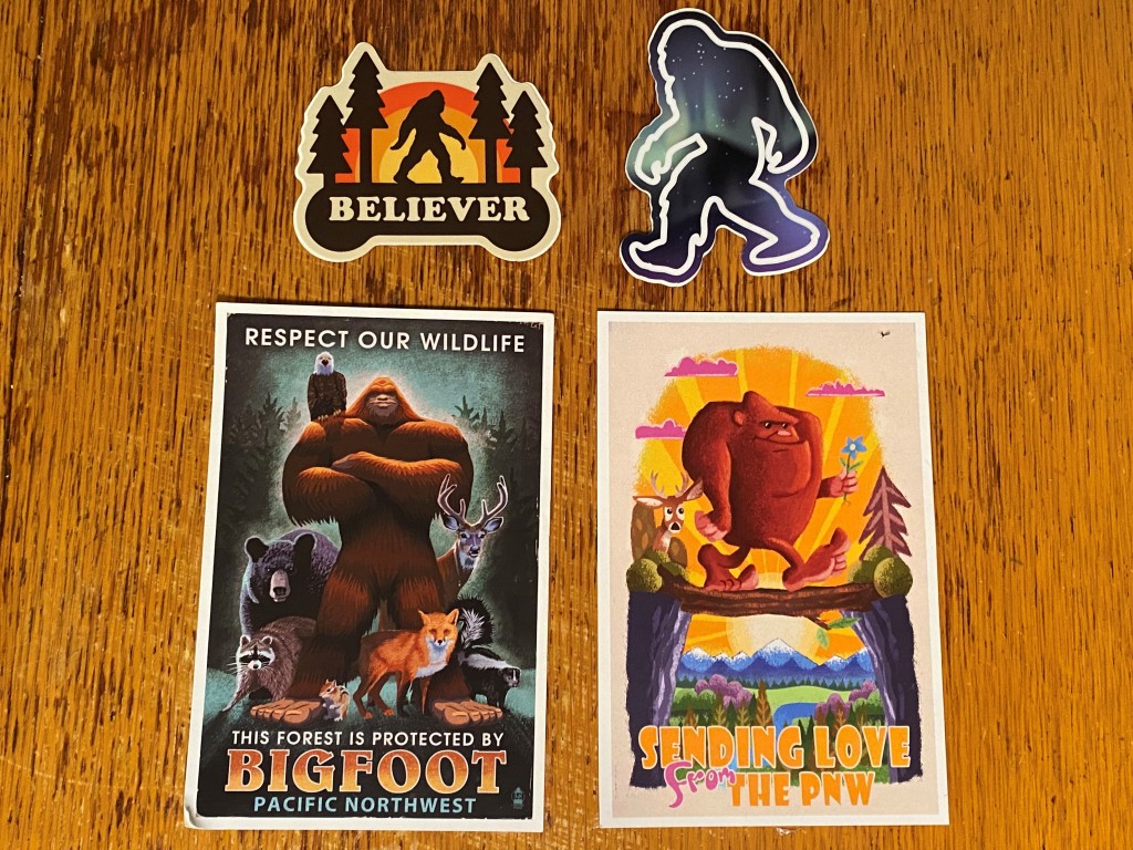 A sticker that says "Believer" with a bigfoot walking between pine trees with a rainbow behind. A sticker of bigfoot with a galaxy inside. A postcard with bigfoot and several wildlife animals that says "Respect our wildlife. This forest is protected by Bigfoot." A postcard with bigfoot and a jackalope walking across a log bridge that says "Sending love from the Pacific Northwest."