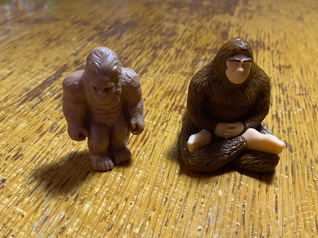 Two plastic Bigfoot figurines. One is standing with ripped abs. The other is sitting in a cross-legged yoga position.